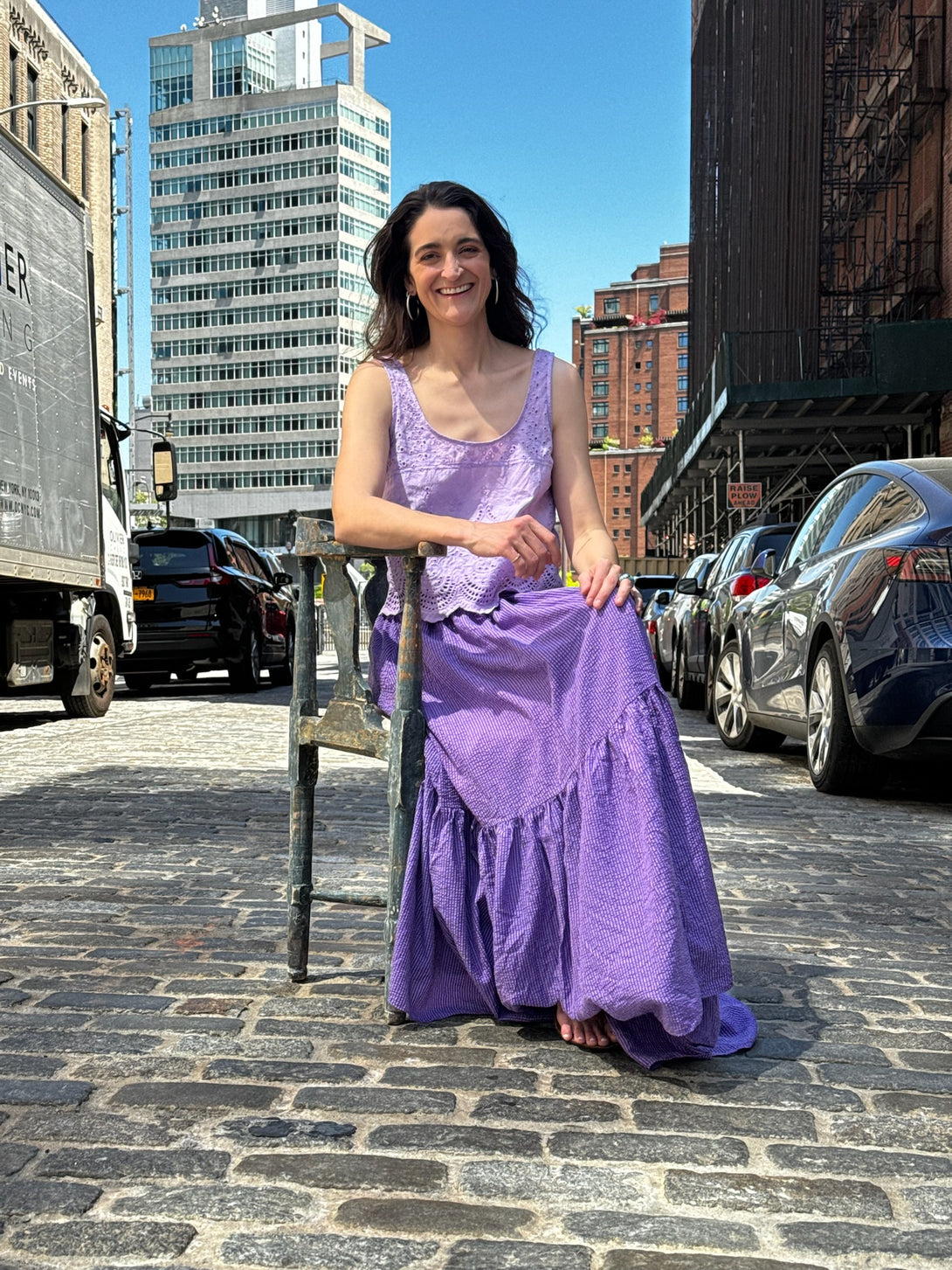 Woman in a purple hand dyed eyelet blouse and peasent skirt standing on a city street with buildings and cars in the background.