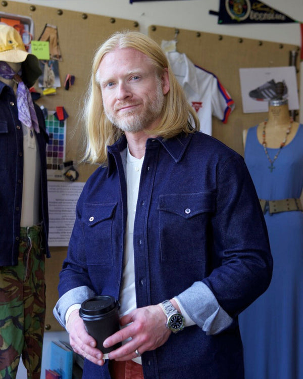 Man with long blonde hair holding a coffee cup in a room with clothing and decor.