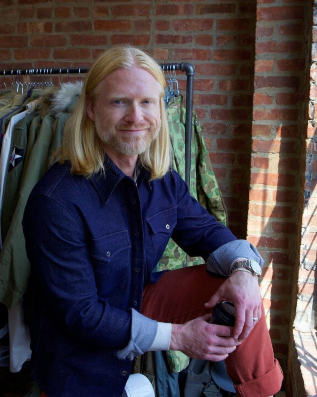 Man with long blonde hair and a beard sitting against a brick wall with coats hanging on racks.